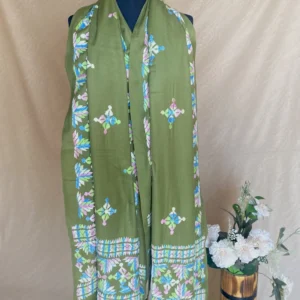 Handwoven sage green cashmere shawl with intricate multicolored embroidery, displayed on a mannequin alongside a vase of white flowers.