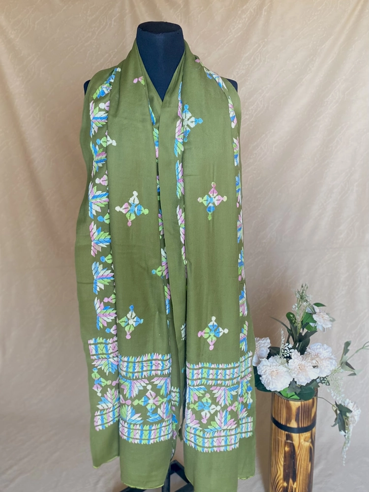 Handwoven sage green cashmere shawl with intricate multicolored embroidery, displayed on a mannequin alongside a vase of white flowers.